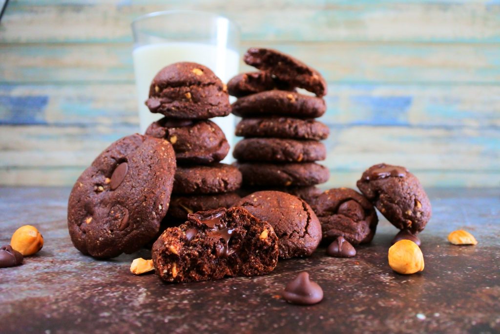 A head on image of two stacks of Double Chocolate Hazelnut cookies with other cookies propped up around the stacks surrounded by chocolate chips and hazelnuts on the tabletop and with a glass of milk in the background
