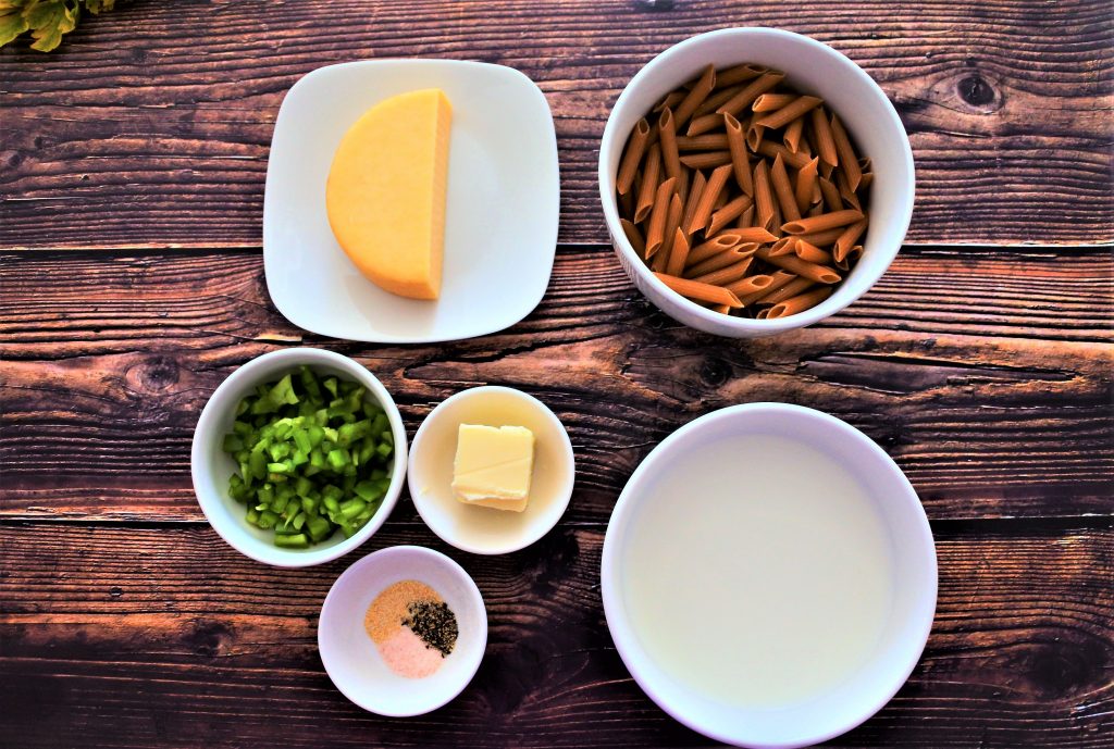 An overhead image of plates and dishes containing ingredients such as: whole grain pasta, milk, smoked Gouda, a pat of butter, salt, black pepper, garlic powder, and diced green bell peppers
