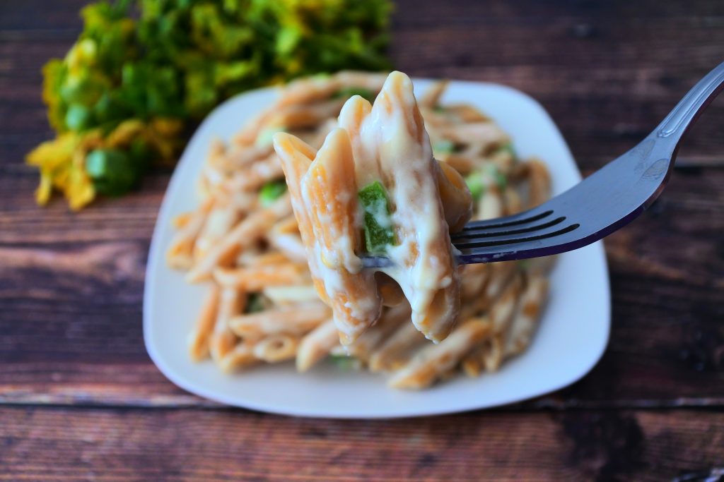 A close up image of a forkful of smoked Gouda mac and cheese with a plate of mac and cheese and a bundle of fresh herbs in the background