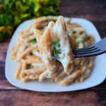A close up image of a forkful of smoked Gouda mac and cheese with a plate of mac and cheese and a bundle of fresh herbs in the background