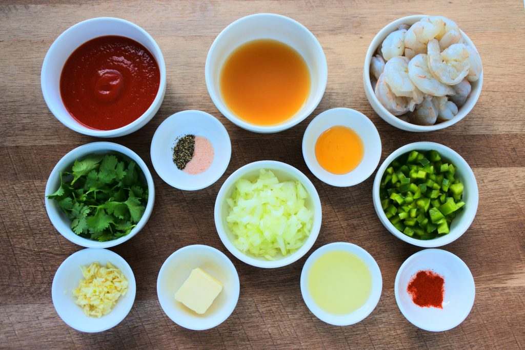 An overhead image of bowls containing ingredients for enchilado de camarones including: shrimps, broth, tomato paste, cilantro, salt and pepper, onions, green bell peppers, garlic, butter, olive oil, smoked paprika and cayenne pepper and honey