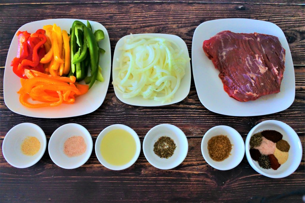 An overhead image of plates and bowls containing the following ingredients: sliced multi-colored bell peppers, sliced onions, a slab of steak, garlic powder, salt, oil, dried oregano, raw sugar, and a bowl of mixed assorted spices