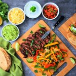 An overhead image of a bamboo board of sliced steak and bell peppers with onions surrounded by bowls of cheese, sour cream, diced tomatoes and guacamole, as well as a prepared fajita on a smaller board, with a stack of flour tortillas in the bottom left and a lemon, avocado and fresh herbs in the top left