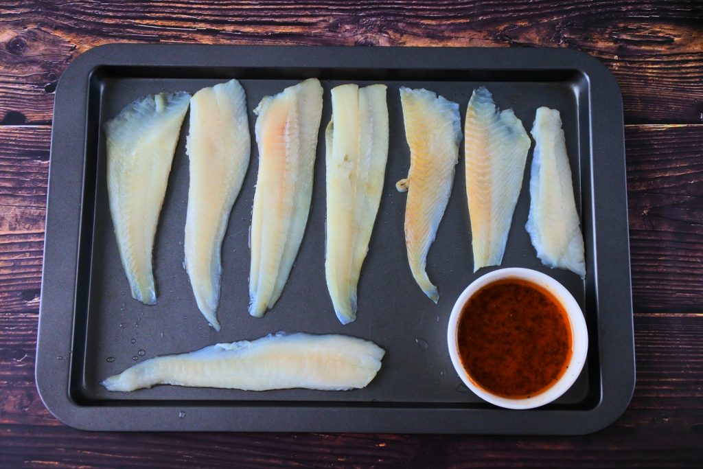 An overhead image of a tray of white fish fillets with a bowl of olive oil based lemon pepper seasoning in the bottom right hand corner of the tray.