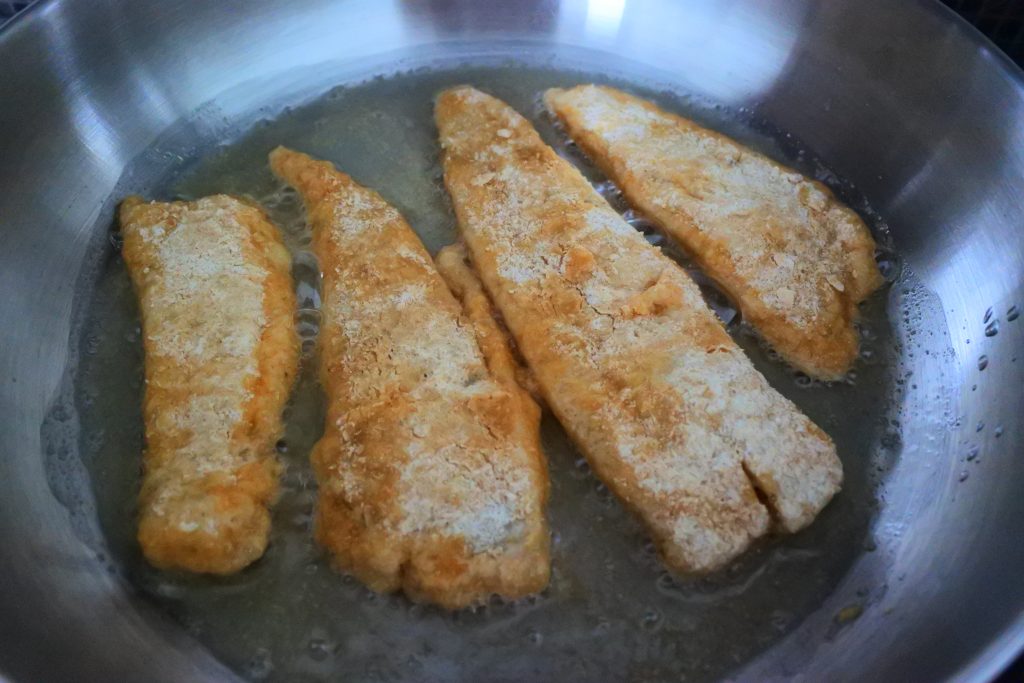 An angled image of four lightly floured fish fillets frying in an olive oil-coated pan.