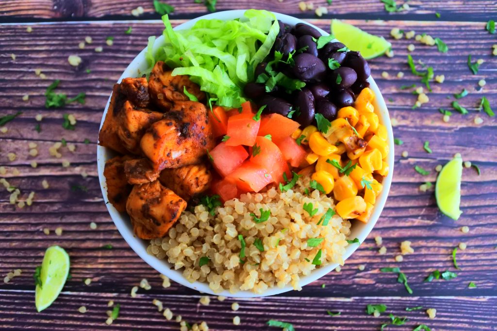 An overhead image of a quinoa power bowl containing fluffy quinoa, chipotle chicken, shredded lettuce, black beans, roasted corn and fresh tomatoes all garnished with finely shredded cilantro and lime wedges