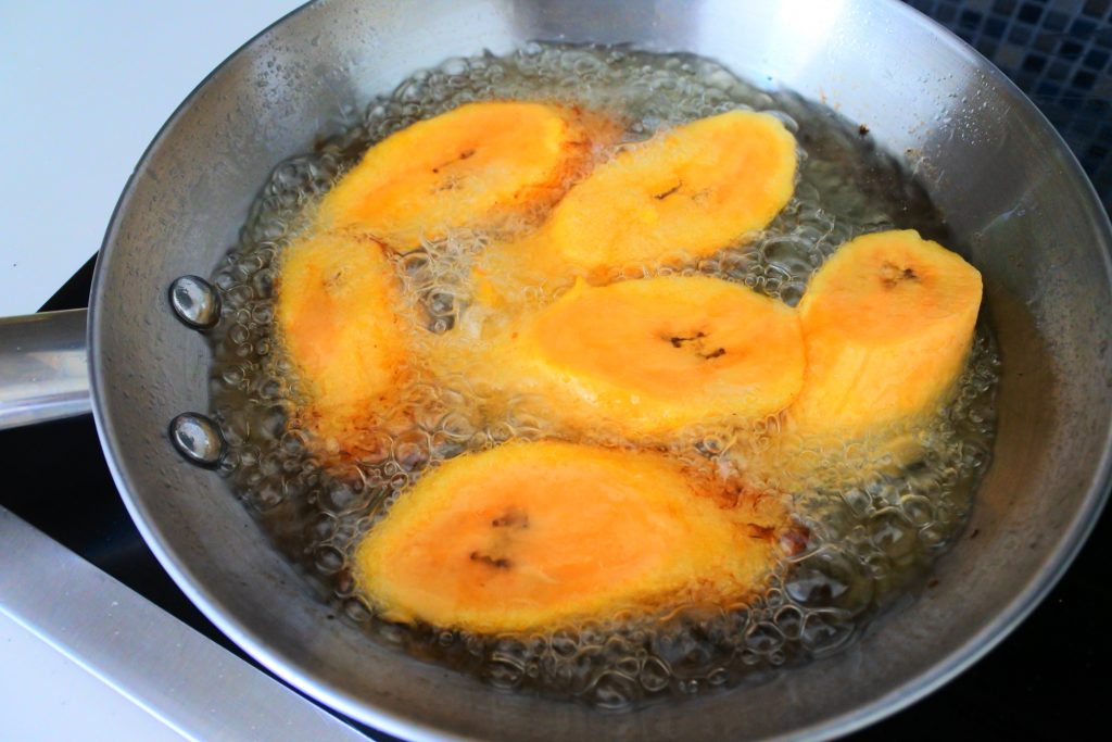 An angled overhead image of slices of plantains in a frying pan being fried in oil.