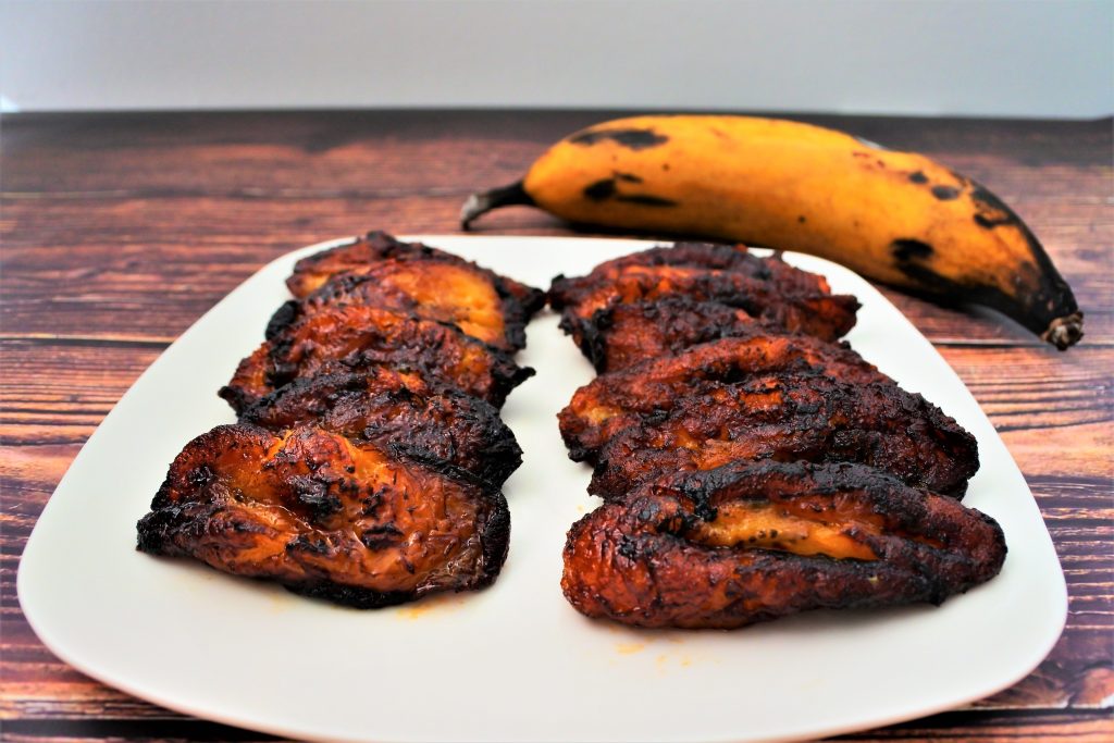 A close up image of slices of fried plantains on a plate with a whole plantain in the background