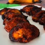 A close up image of slices of fried plantains on a plate with a whole plantain in the background