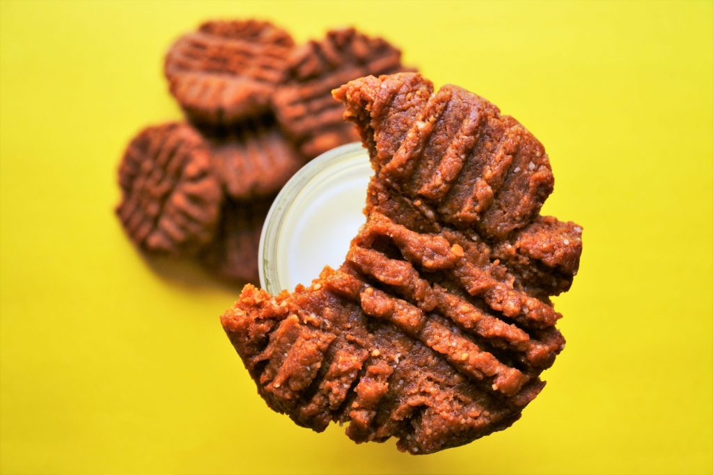 An overhead image of a peanut butter cookie with a bite taken out of it balancing on the neck of a bottle of milk with a pile of peanut butter cookies below on a yellow surface