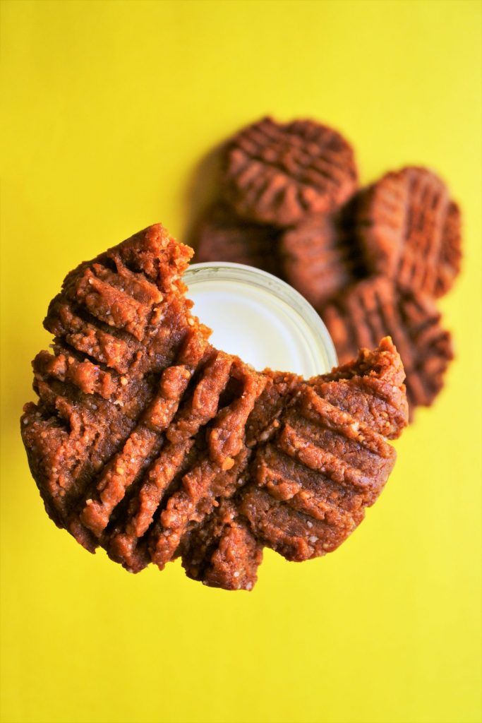 An overhead image of a peanut butter cookie with a bite taken out of it balancing on the neck of a bottle of milk with a pile of peanut butter cookies below on a yellow surface