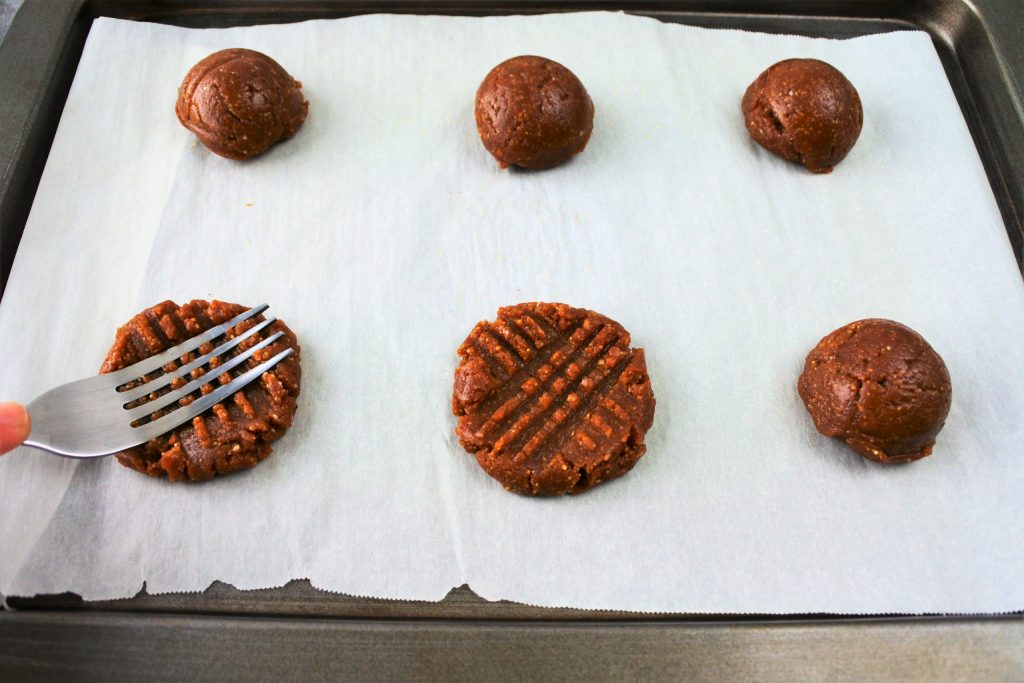 An angled image of a parchment lined tray with portions of peanut butter cookie dough on it. One round of dough has been pressed with a cross hatch mark while another is being pressed down with the flat tines of a fork