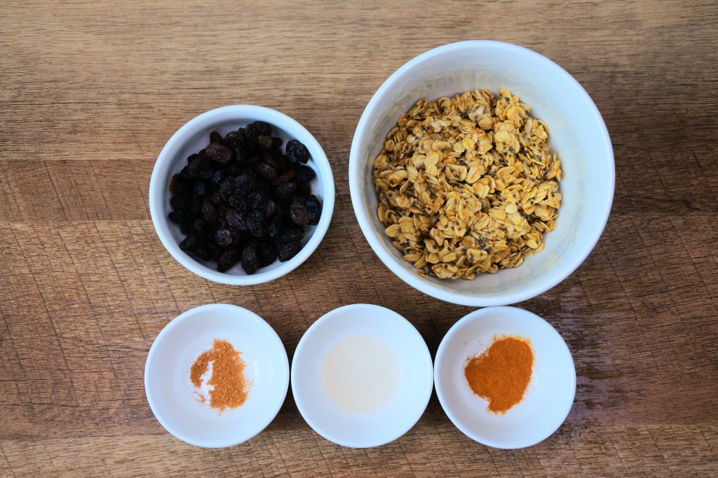 An overhead image of bowls containing ingredients such as nutmeg, cinnamon, vanilla extract and raisins.