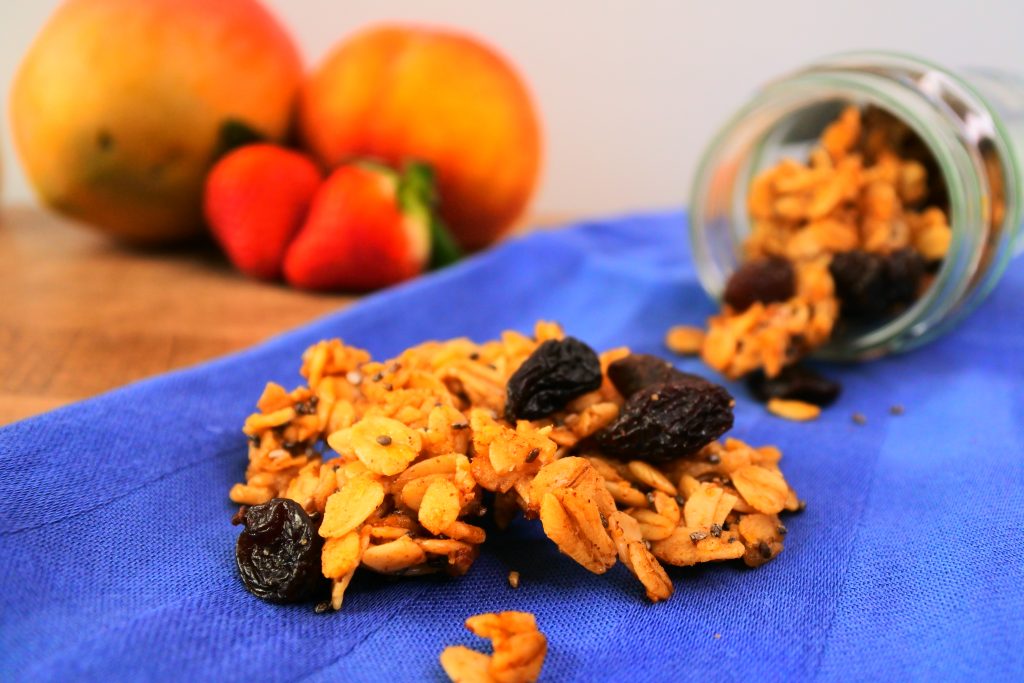 A close up image of a clump of oatmeal raisin granola on a blue cloth with an overturned jar of granola and fresh fruit in the background