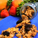 A head on image of an overturned jar of oatmeal raisin granola with the granola spilling out onto a blue cloth with fresh fruit in the background