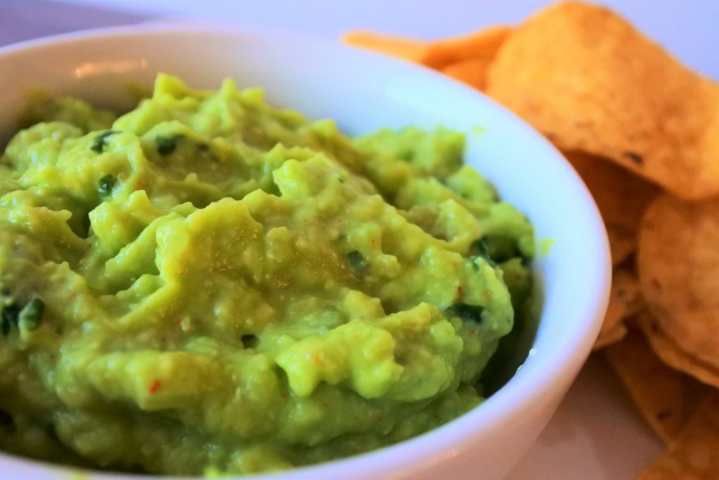 An angled head on image of a bowl of guacamole with yellow corn tortilla chips in the background