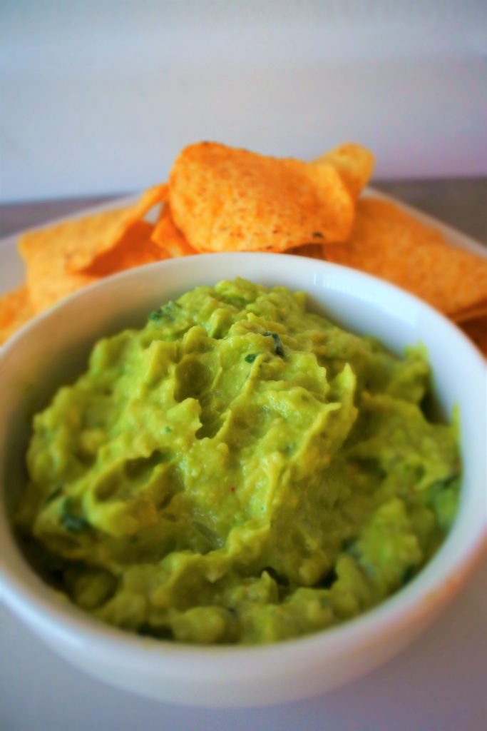 A tilted angle image of a bowl of guacamole with yellow corn tortilla chips in the background
