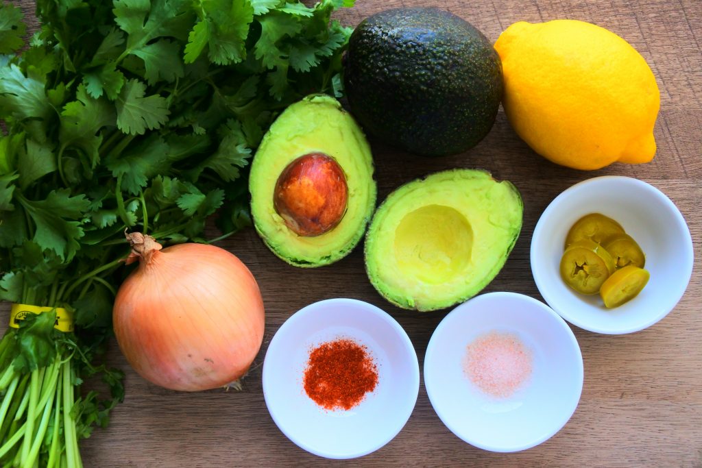 An overhead image of an assortment of ingredients used for guacamole including fresh cilantro, onion, avocados, lemons, sliced jalapenos, salt and cayenne pepper.