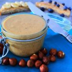 A close up image of a jar of homemade hazelnut butter on a blue cloth napkin surrounded by hazelnuts with a knife leaning against the jar and pieces of toast covered with hazelnut butter and fruit in the background