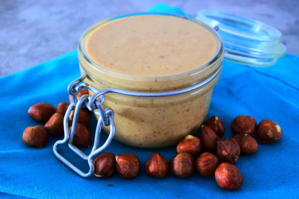 A close up image of a jar of homemade hazelnut butter on a blue cloth napkin surrounded by hazelnuts
