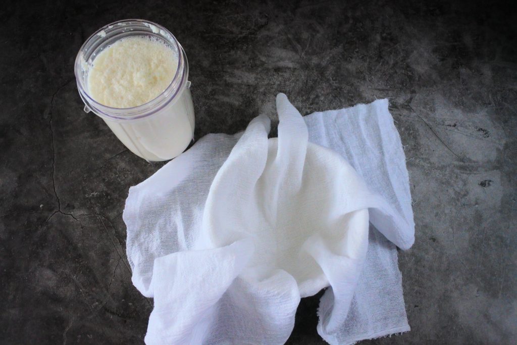 An overhead image of blended coconut and water in a blender jug next to a bowl with a cheese cloth in it.