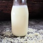 Head on image of a bottle of fresh coconut milk surrounded by shredded coconut.