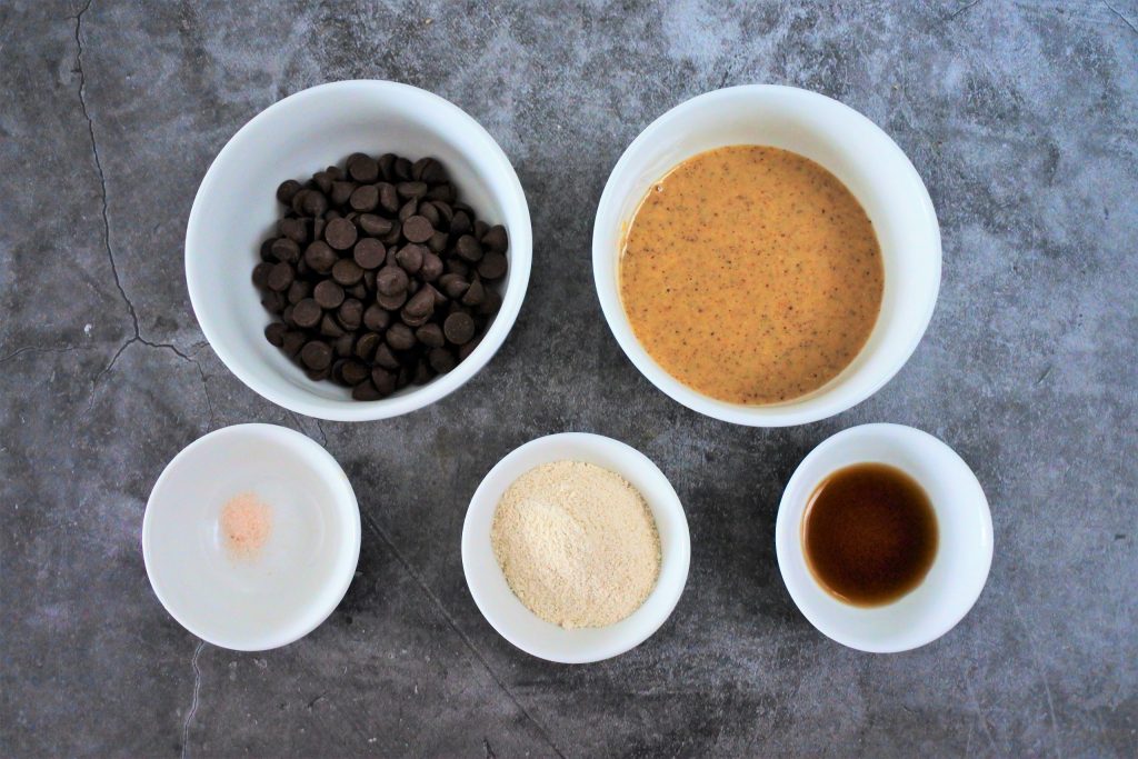 An overhead image of bowls of ingredients for a homemade chocolate hazelnut spread including dark chocolate chips, homemade hazelnut butter, vanilla extract, finely ground raw cane sugar, and a pinch of salt