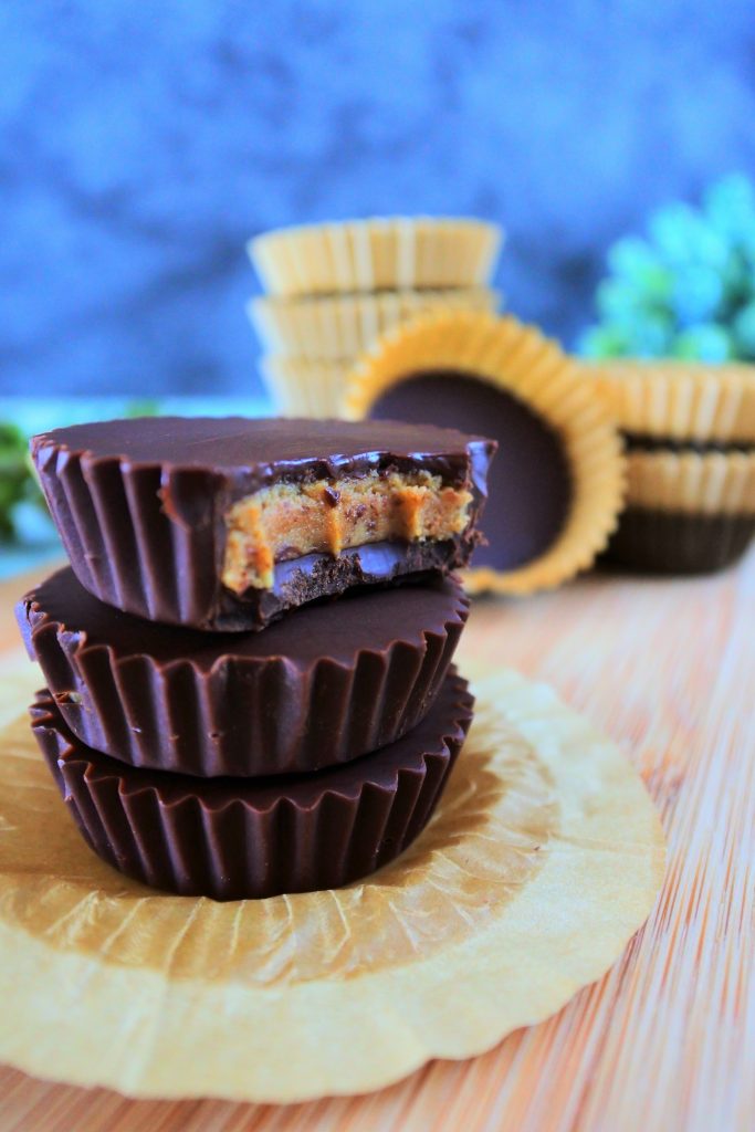 A head on close up image of a stack of three peanut butter cups with a bite taken out of the top one with paper lined peanut butter cups in the background