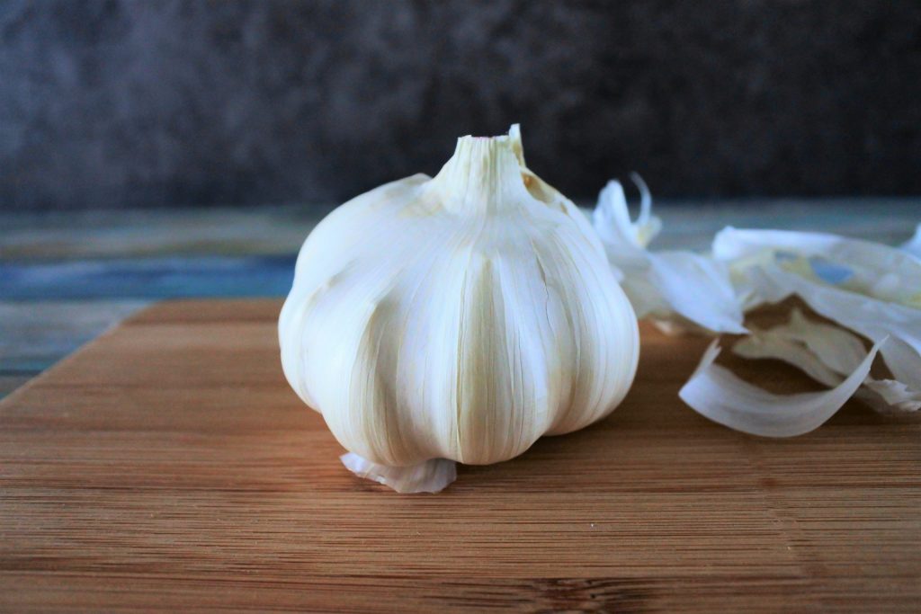 A head on image of a head of garlic on a wooden board with layers of outer skin peeled off and on the right side of the board