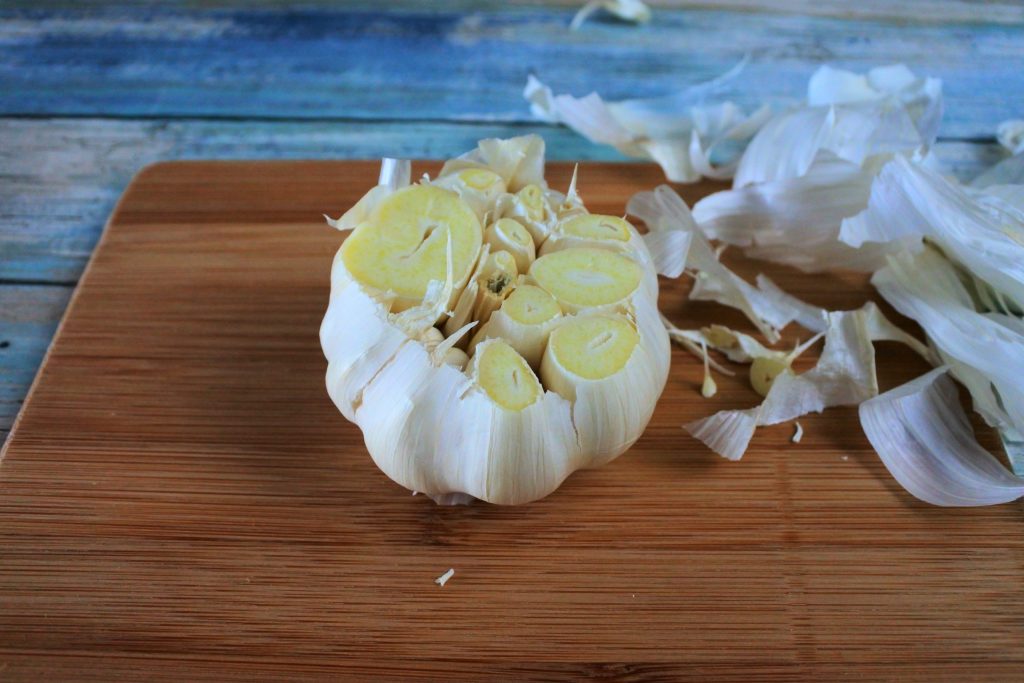 An angled image of a cut head of garlic on a wooden board with pieces of the papery skin to the right side of it