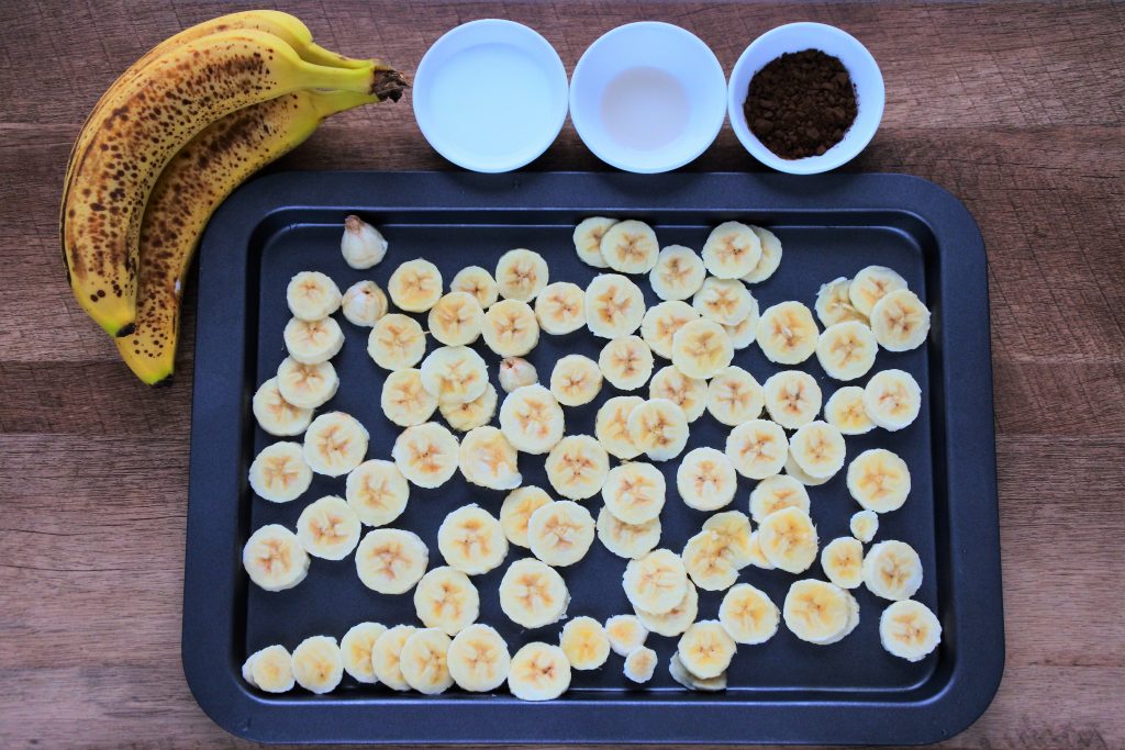 An overhead image of a tray of sliced frozen bananas with a whole banana in the top left corner and three dishes containing coconut milk, vanilla extract and cocoa powder