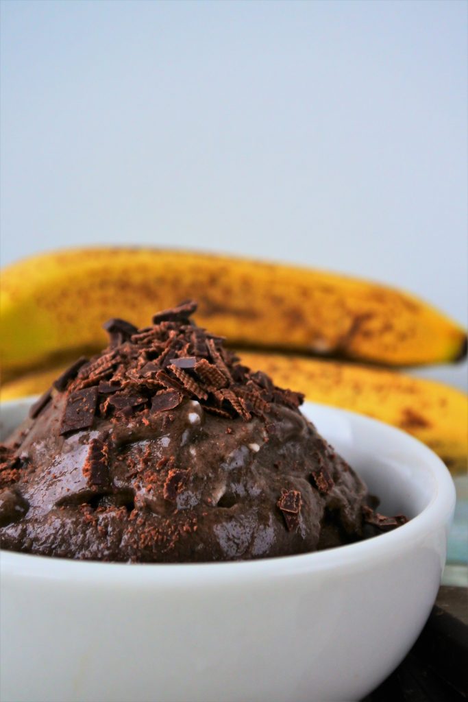 A close up image of a chocolate ice cream topped with shaved chocolate with some whole bananas in the background