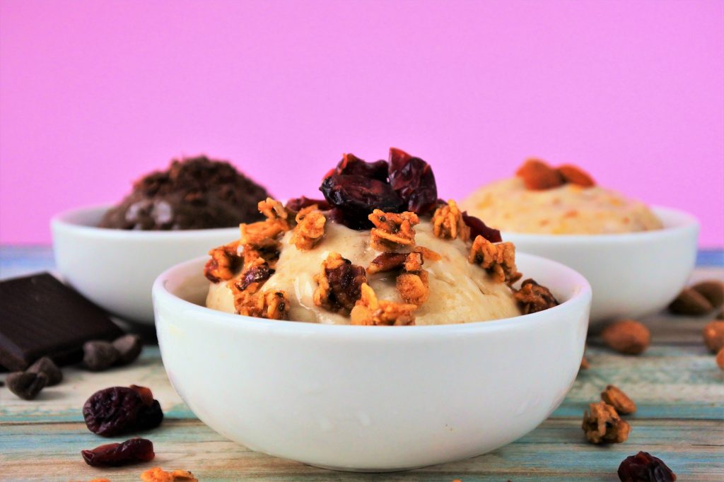 An head on image of three bowls of ice cream. One containing chocolate ice cream topped with and surrounded by chocolate, the other containing an amaretto ice cream topped with and surrounded by almonds and the third containing a base banana and vanilla ice cream topped with and surrounded by granola and dried cranberries