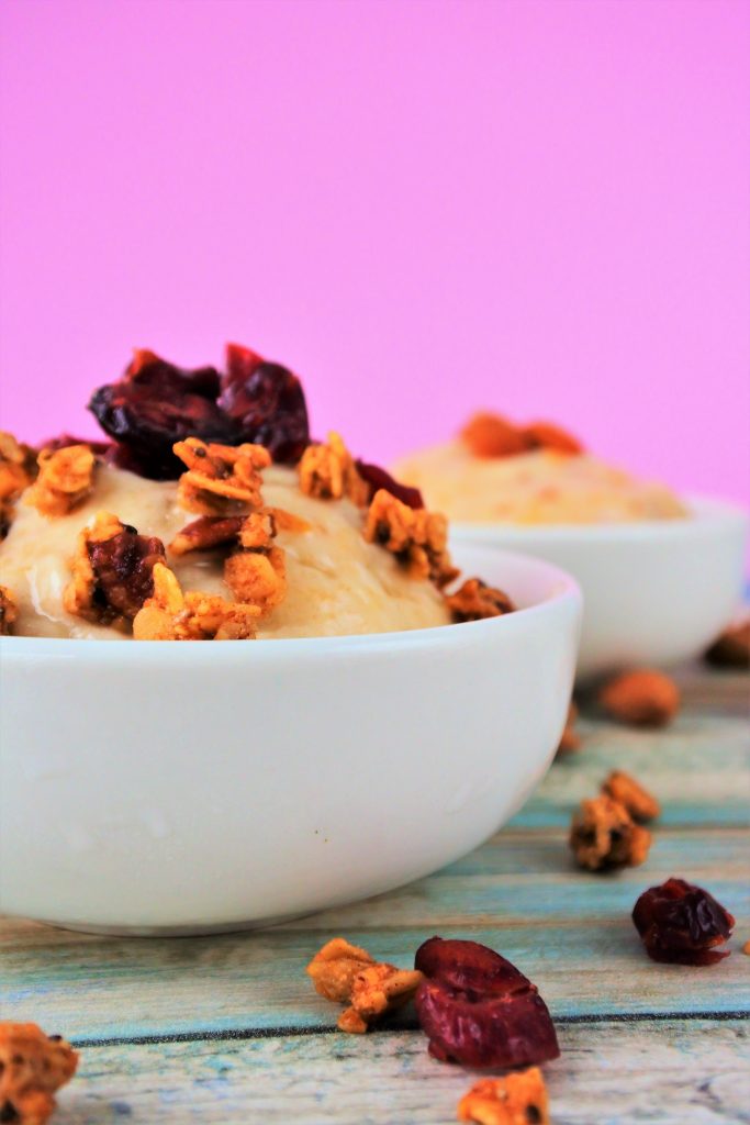 a close up image of a bowl of banana ice cream topped with and surrounded by homemade granola and dried cranberries.