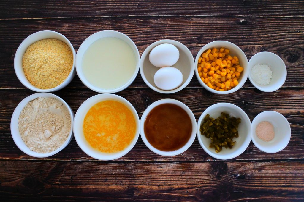 An overhead image of bowls containing ingredients for Jalapeno cornbread including corn meal, whole wheat pastry flour, buttermilk, melted butter, eggs, honey, sweet corn kernels, diced jalapenos, baking powder and salt.