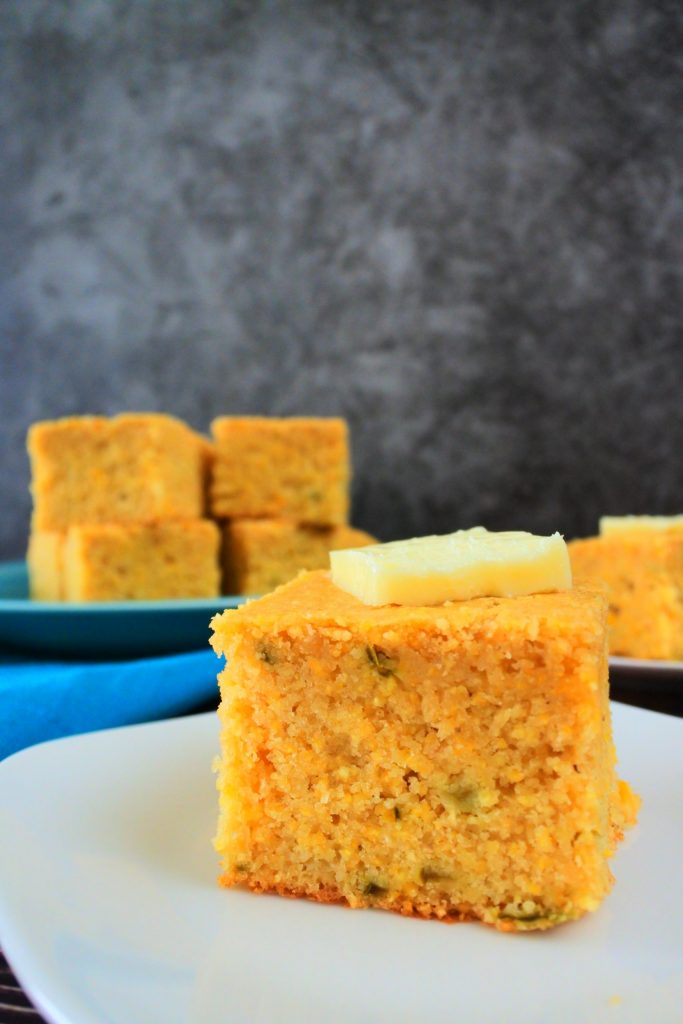 A head on image of a piece of jalapeno cornbread on a plate topped with a pat of butter with a plate of cornbread pieces in the background