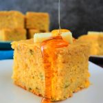 A head on image of a piece of jalapeno cornbread on a plate topped with a pat of butter and a drizzle of honey that spilled over the sides with a plate of cornbread pieces in the background