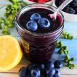 An angled image of a jar of blueberry jam with fresh blueberries on top and a spoon in it surrounded by fresh fruit including blueberries, raspberries, blackberries and a half cut lemon