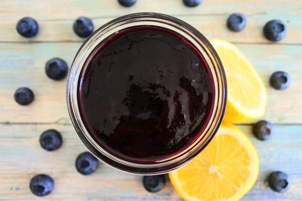 An overhead image of blueberry jam in a jar with fresh blueberries and cut lemons at the base
