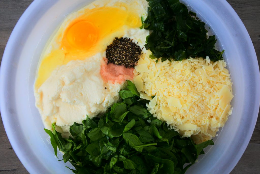 An overhead image of a bowl of ingredients for a spinach and ricotta mixture including: ricotta, spinach, Parmesan cheese, basil, salt, black pepper and an egg