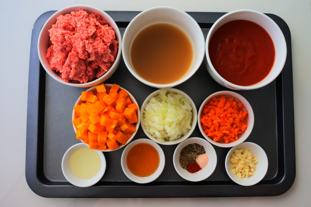 An overhead image of a tray of ingredients for the lasagna's meat sauce including: Meat, broth, tomato sauce, butternut squash, onions, carrots, olive oil, honey, salt, pepper, smoked paprika, dried basil, oregano, rosemary and diced garlic