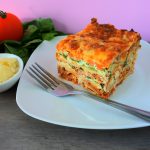 A head on image of a slice of lasagna on a plate with a fork and fresh spinach, tomatoes and a small dish of Parmesan on the side