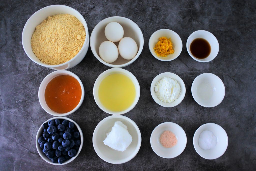An overhead image of ingredients for a lemon-blueberry almond flour cake including: blanched almond flour, eggs, honey, fresh blueberries, lemon juice, coconut oil, lemon zest, vanilla extract, potato starch, lemon extract, Himalayan salt and baking soda