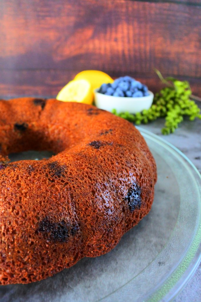 A close up of a lemon blueberry bundt cake on a glass plate with fresh lemons and blueberries in the background