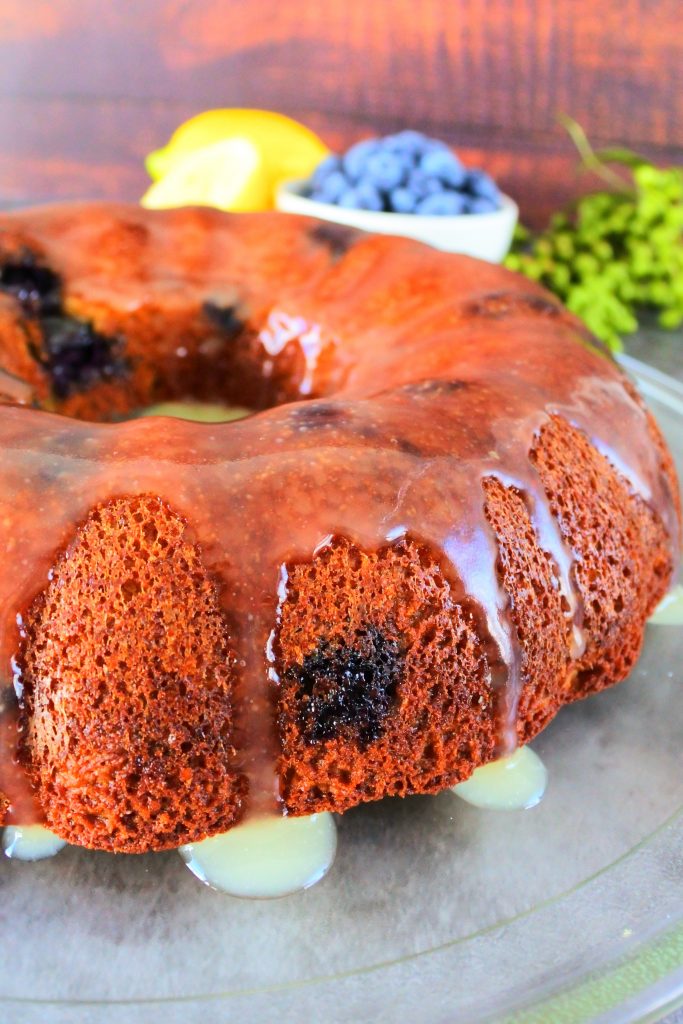 A close up of a glazed lemon blueberry bundt cake on a glass plate with fresh lemons and blueberries in the background