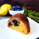 A close up image of a slice of glazed lemon blueberry bundt cake on a plate and fresh lemon and blueberries in the background
