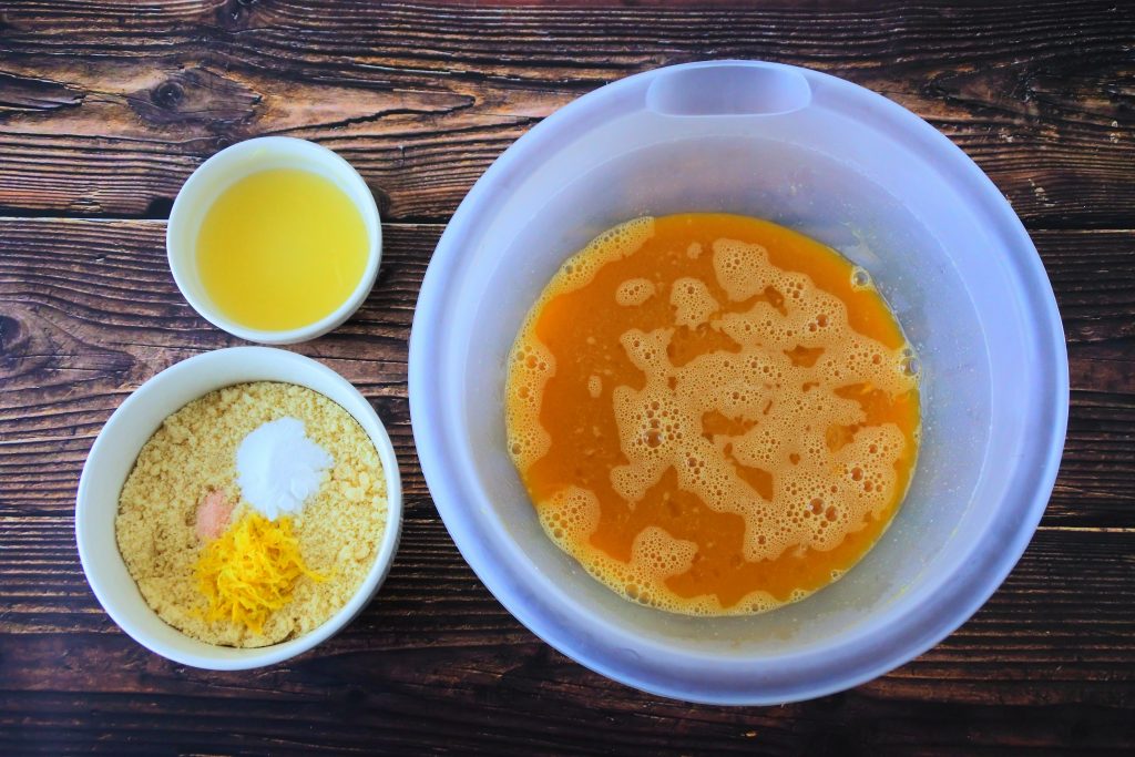 An overhead image of three bowls; one with eggs, honey, lemon extract and vanilla extract, one with lemon juice and one with almond flour, lemon zest, salt and baking soda and powder mix.