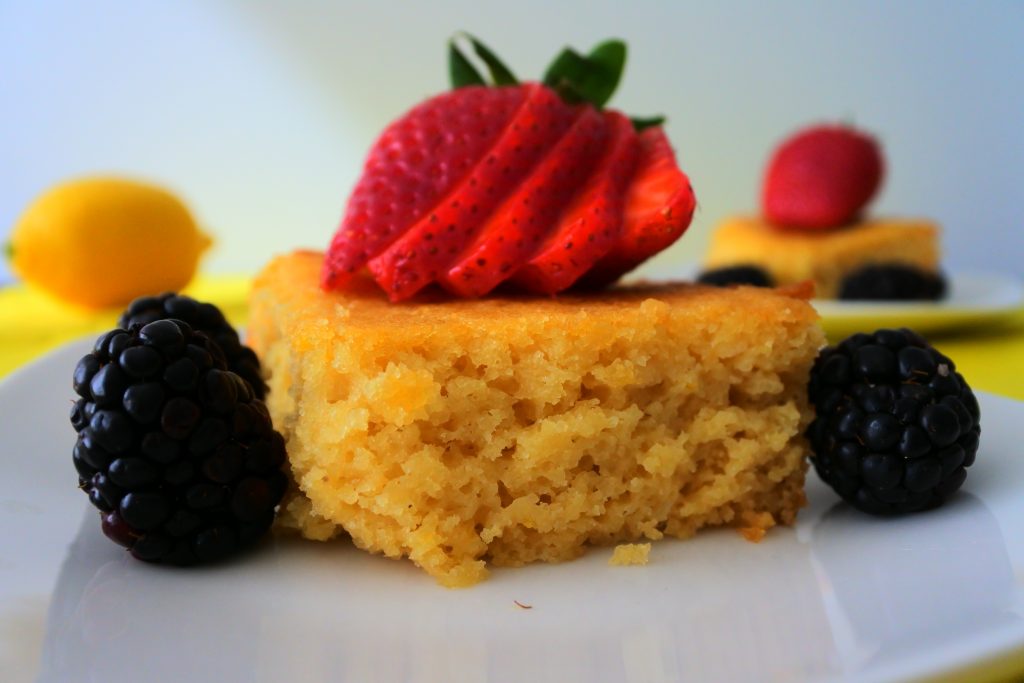 A close up image of a square of lemon cake topped with a sliced strawberry and surrounded by blackberries