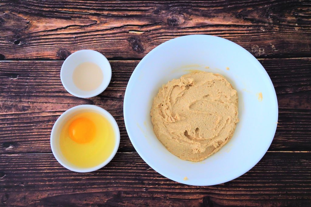 An overhead image of a bowl of creamed egg and sugar with an egg and vanilla extract in bowls on the side.