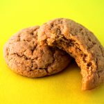 Close up image of two lemon sugar cookies with a cracked tops and a bite taken out of one on a yellow background
